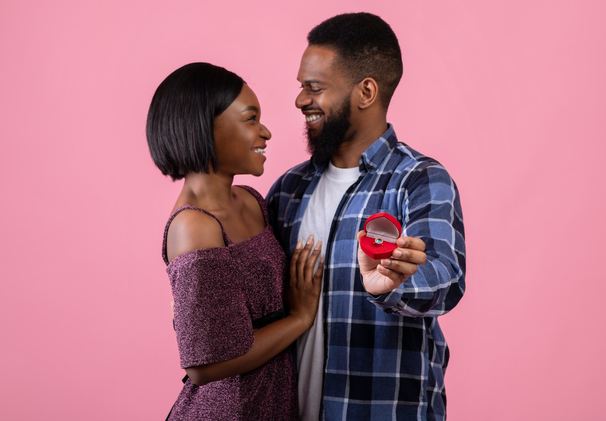 Affectionate black couple hugging and holding box with engagement ring on pink background, selective